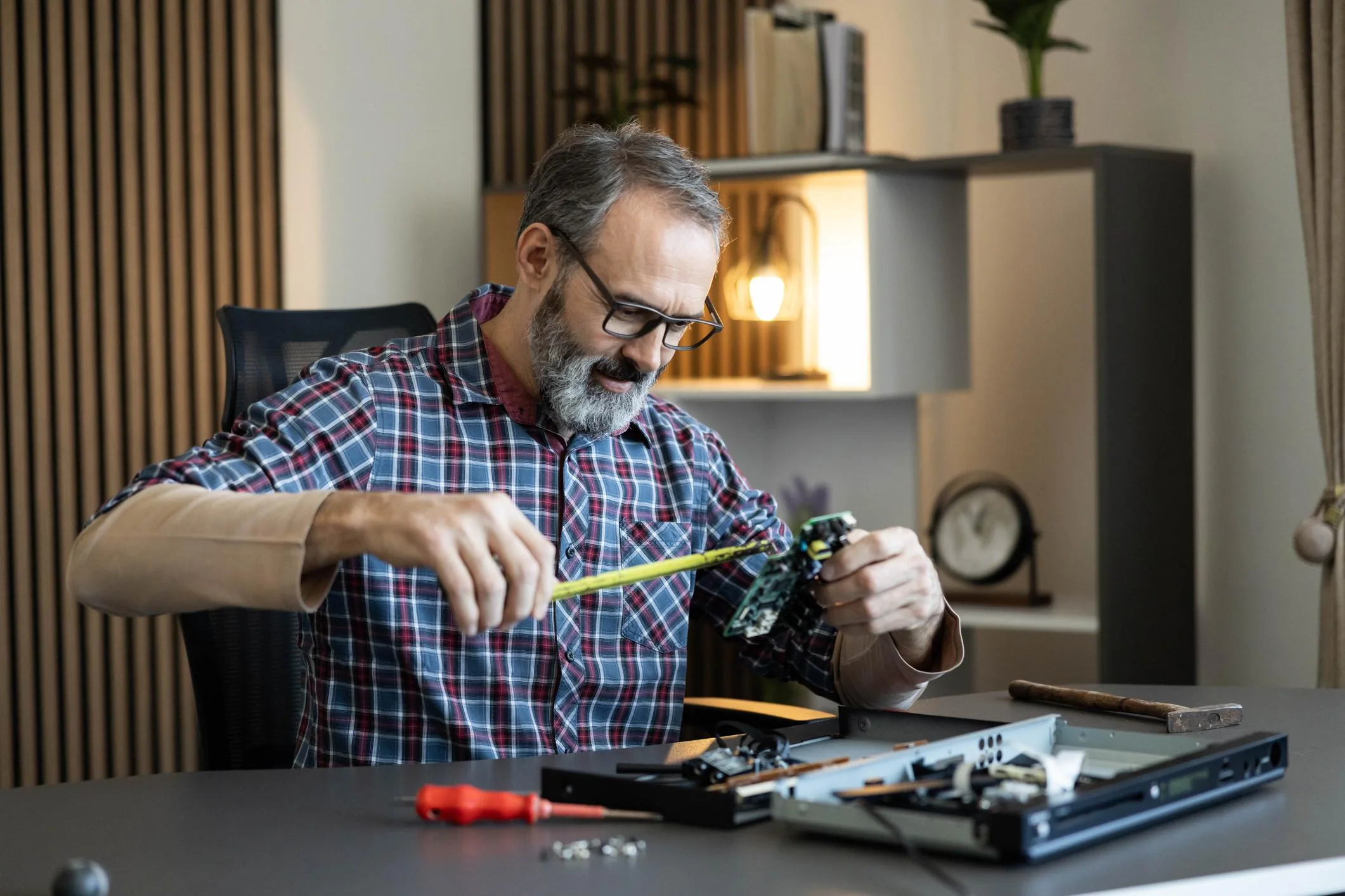 Technician at a tidy home workbench servicing a flat-panel monitor mainboard with a screwdriver under warm lamp light