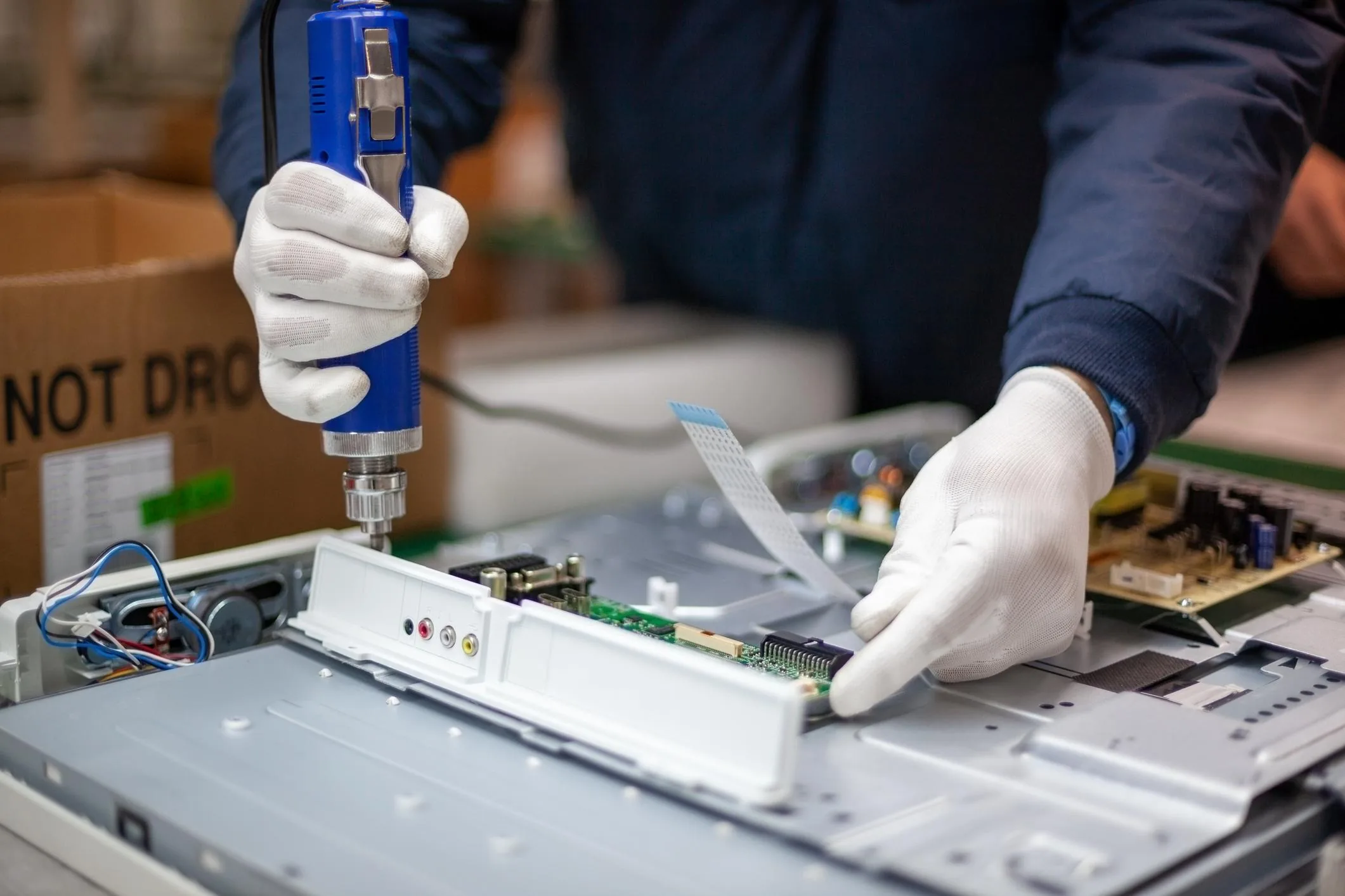 White anti-static gloved hands using a cordless screwdriver to lift a computer monitor mainboard from its chassis