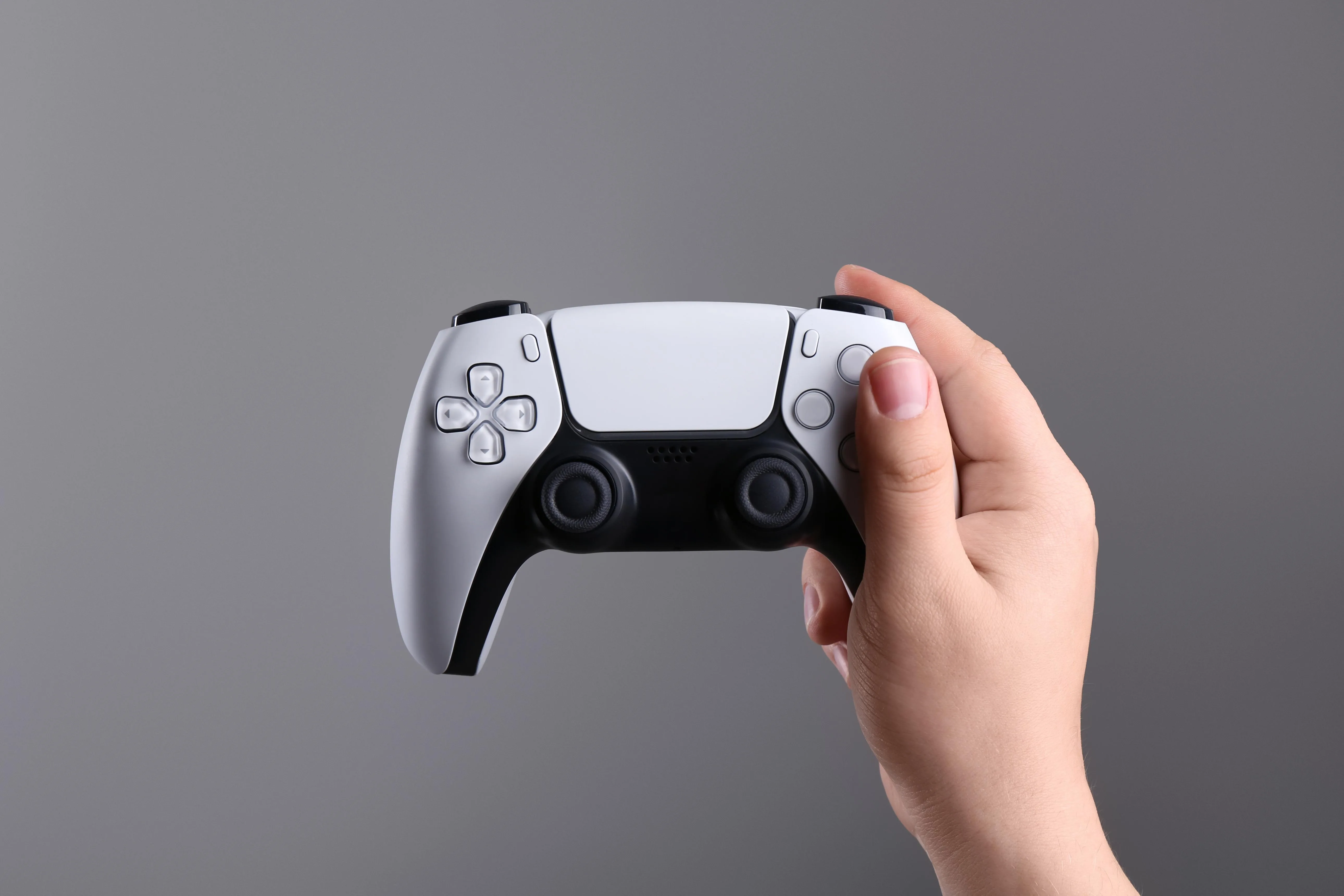 Hand holding a white PlayStation 5 DualSense controller against a clean grey studio backdrop