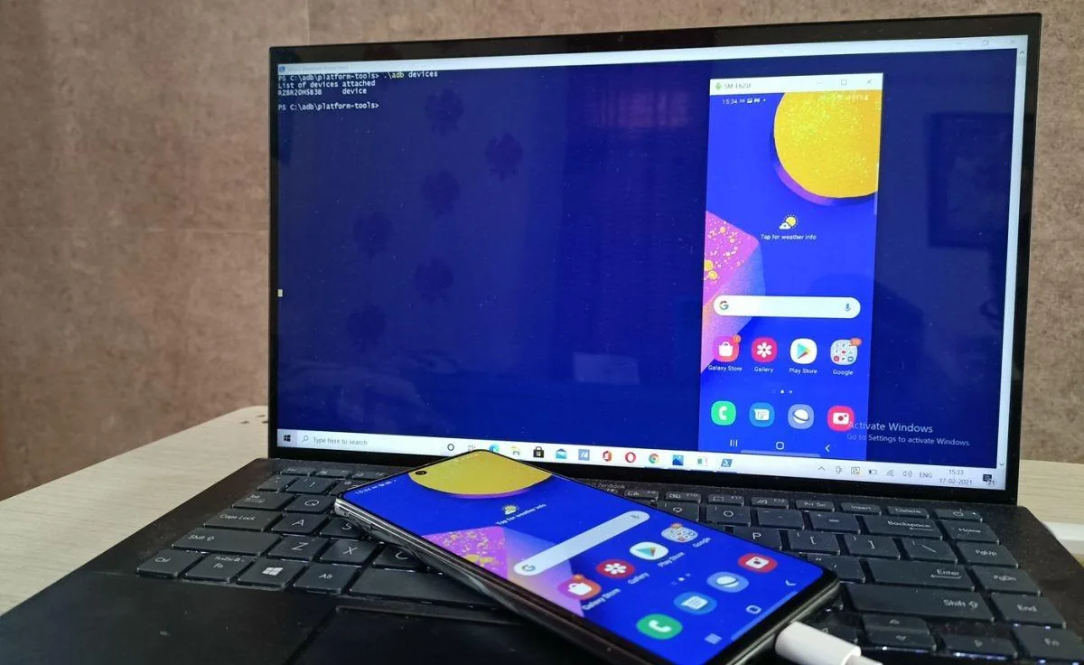 Samsung Galaxy phone resting on a laptop keyboard with the laptop screen mirroring the phone's home screen via an ADB devices command in PowerShell