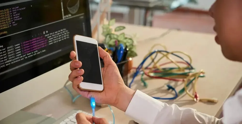 A person holding a white smartphone connected by a glowing blue USB cable to a desktop computer displaying lines of debug log output