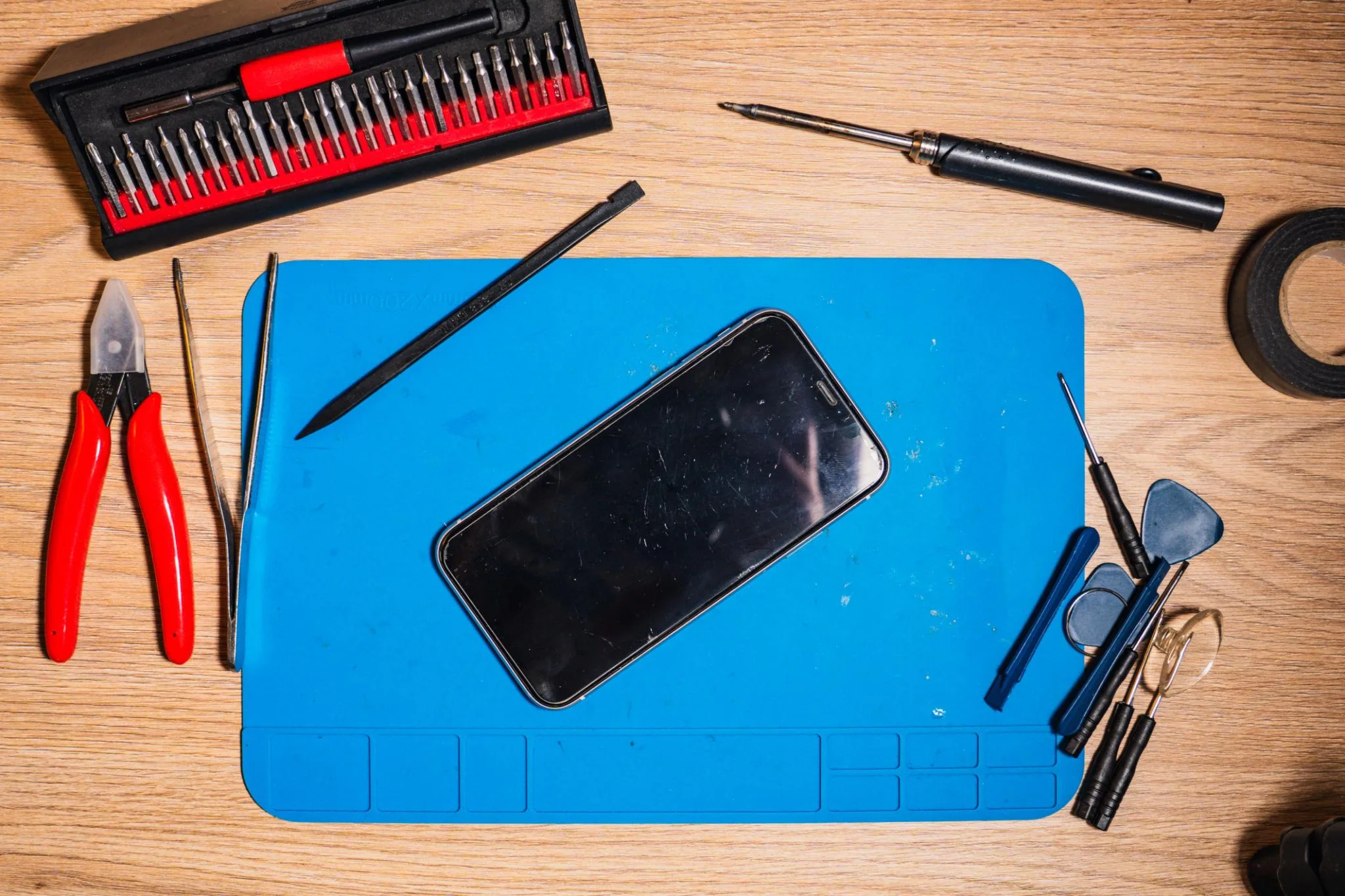 Top-down flat lay of a smartphone resting on a blue silicone repair mat surrounded by a complete precision screwdriver kit, plastic spudgers, tweezers and red flush cutters on a wooden surface